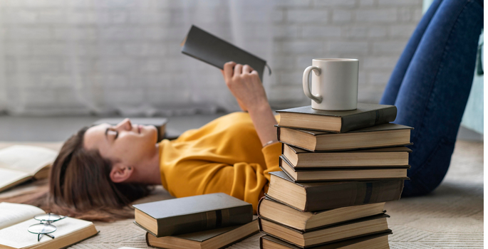A teenage girl lies on her back reading behind a stack of books and a coffee mug.