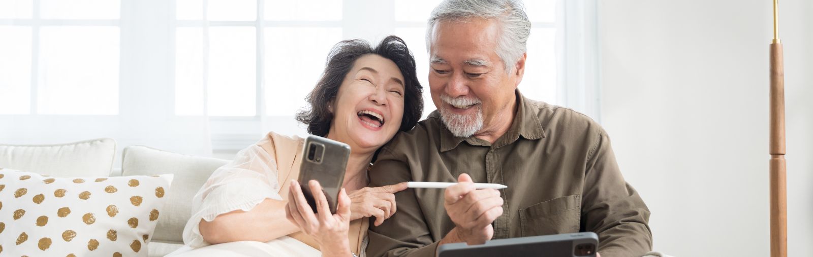 Two older adults of Asian descent laughing at a smartphone and tablet