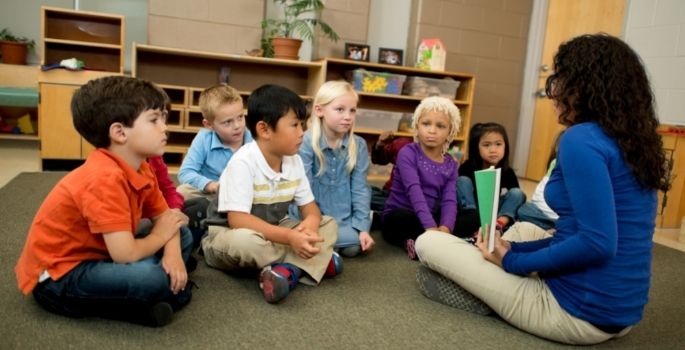 A group of children sitting on the floor listening to a woman reading a book