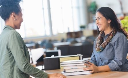 A smiling woman checks out books for another woman.