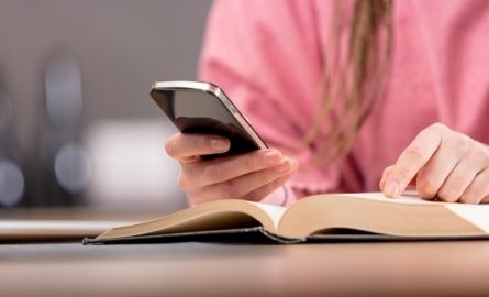 A woman with a book on a table checks her cell phone